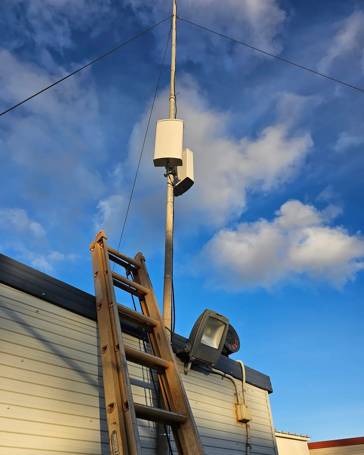 Two white panel antennas on a guyed mast above a site building, with a ladder against the wall
